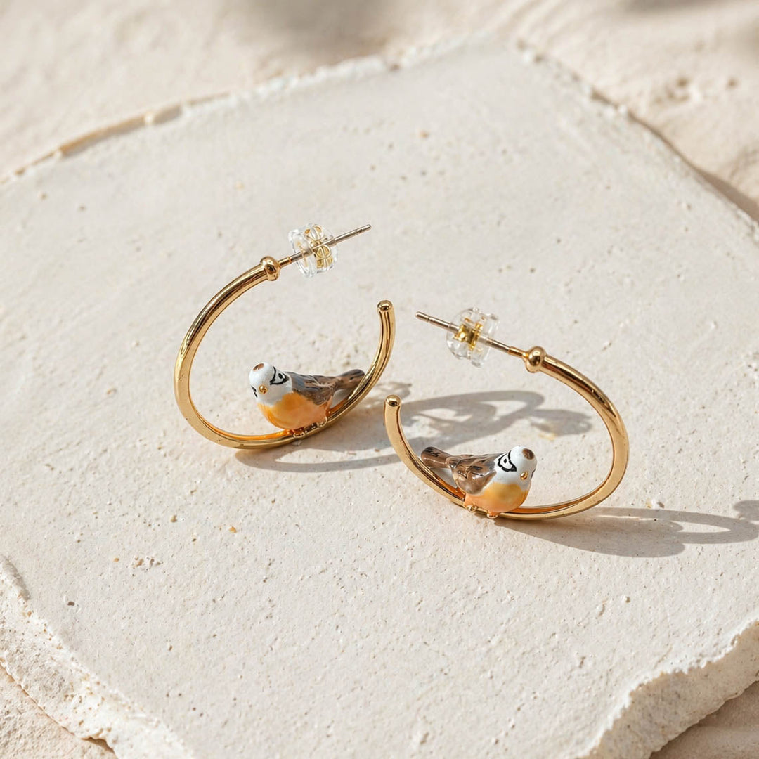 Gold hoop earrings with bird designs on a textured beige surface