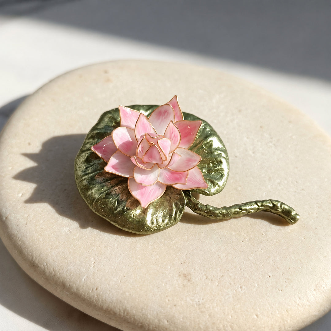 Pink lotus floral brooch with layered petals and green enamel lily pad on a sunlit cream stone surface.
