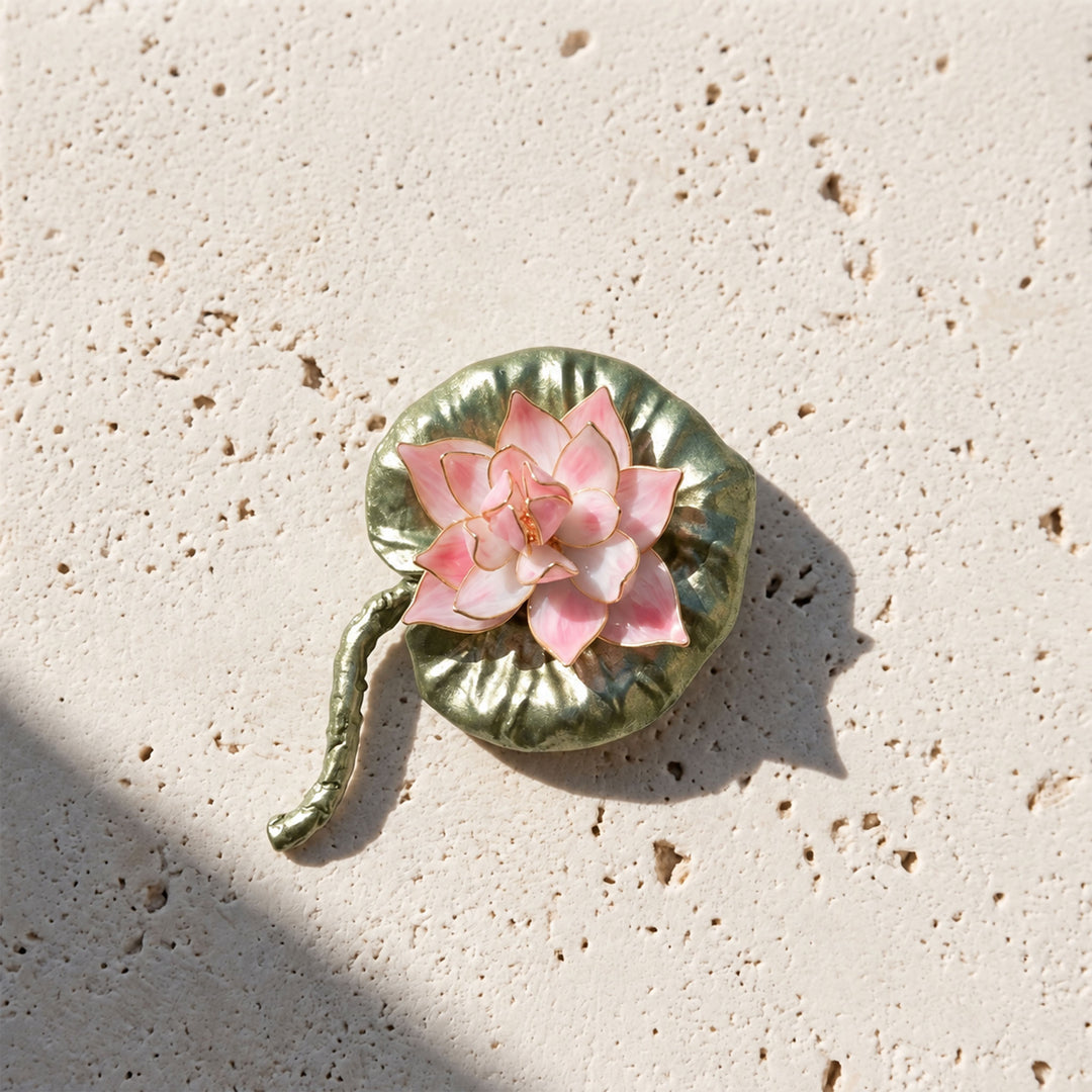 Handcrafted pink lotus brooch with textured green enamel leaf and stem on pale stone in direct sunlight.