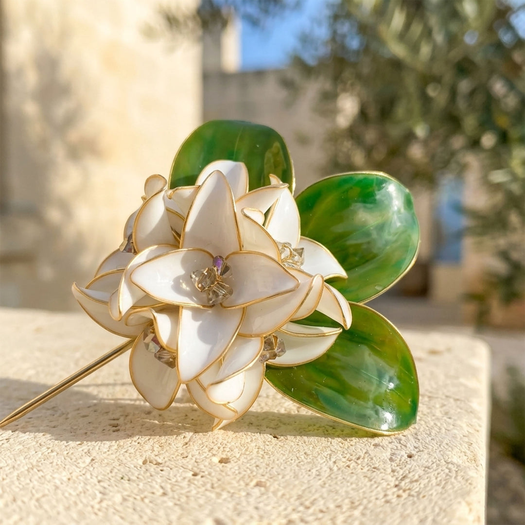 Close-up of a handcrafted white gardenia flower brooch with sculptural green leaves and gold-tone metal outlines, resting on a textured beige stone surface with a blurred outdoor background.