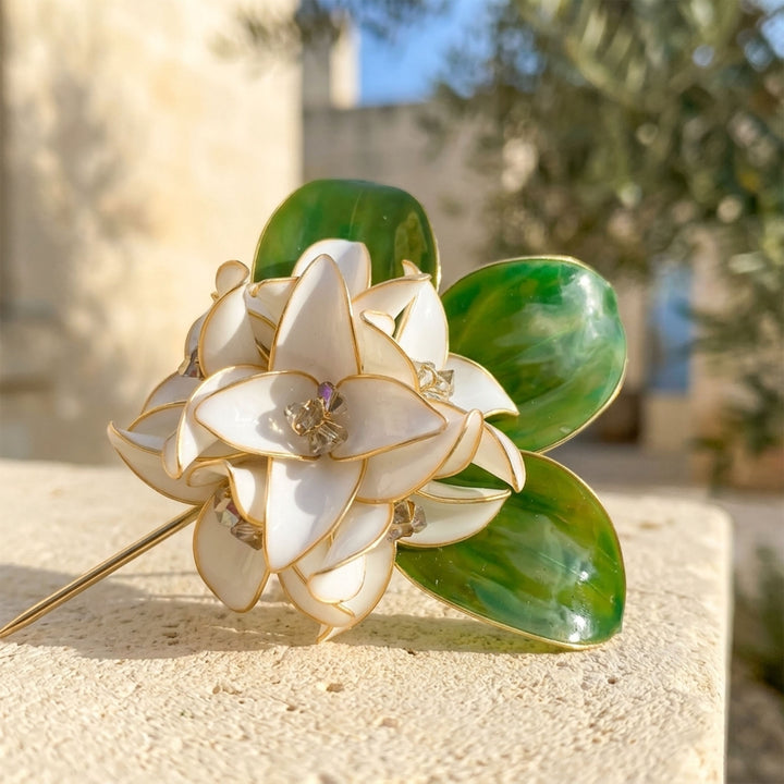 Close-up of a handcrafted white gardenia flower brooch with sculptural green leaves and gold-tone metal outlines, resting on a textured beige stone surface with a blurred outdoor background.
