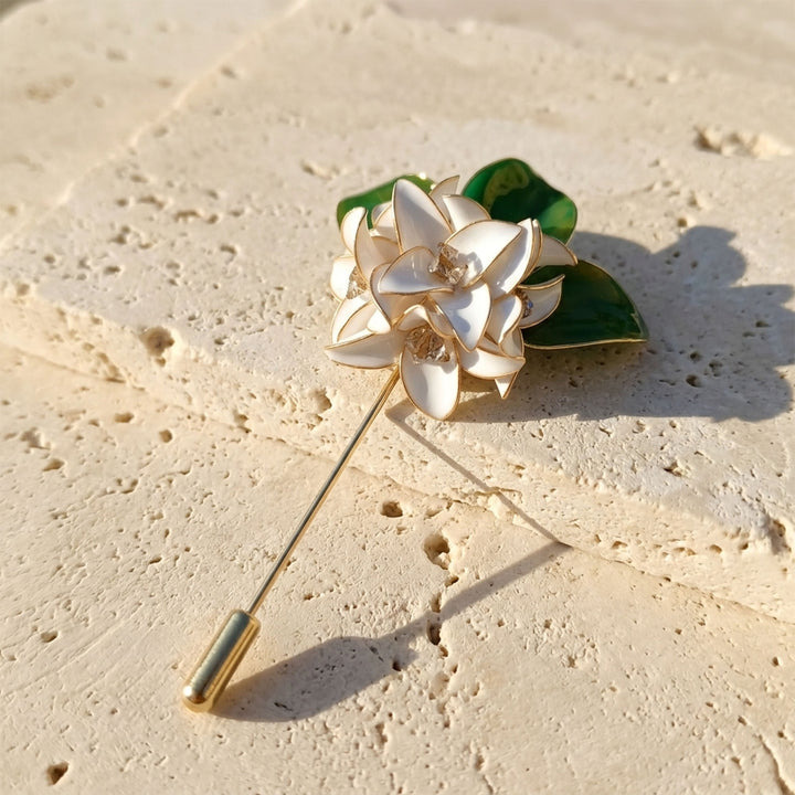 Handcrafted white gardenia floral pin brooch with green leaves and glass details, resting on a textured beige stone surface.