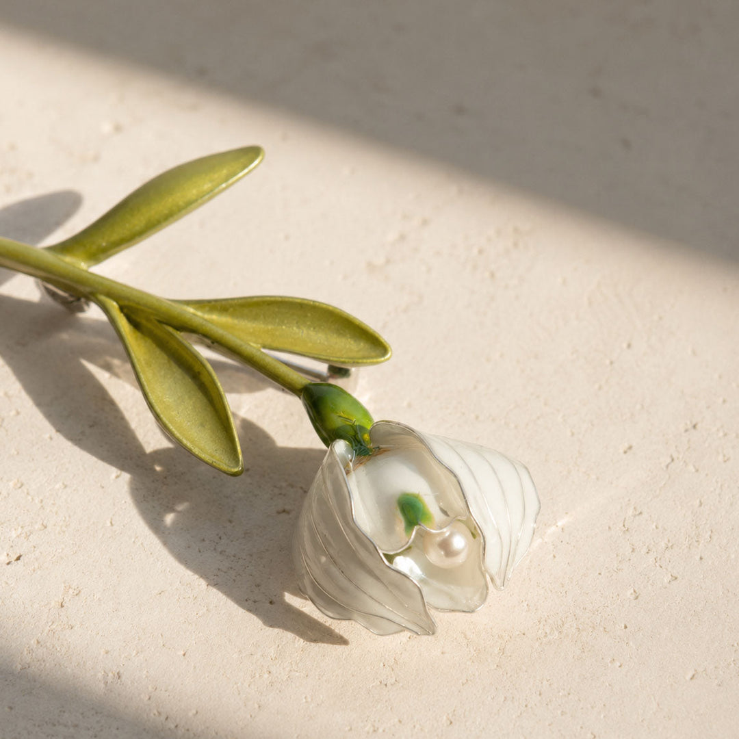Lily of the valley pearl brooch with hand-painted enamel flower resting on a light stone surface.