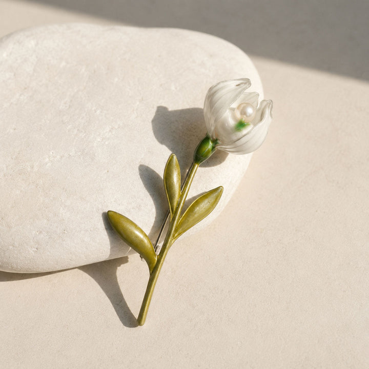 Handcrafted lily of the valley pearl brooch with white enamel bloom and green leaves on a smooth white stone.