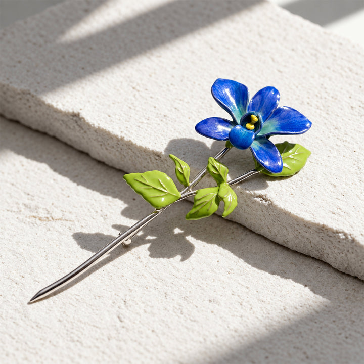 Blue orchid floral pin brooch with hand-painted enamel petals and green leaves on a sunlit pale stone surface.