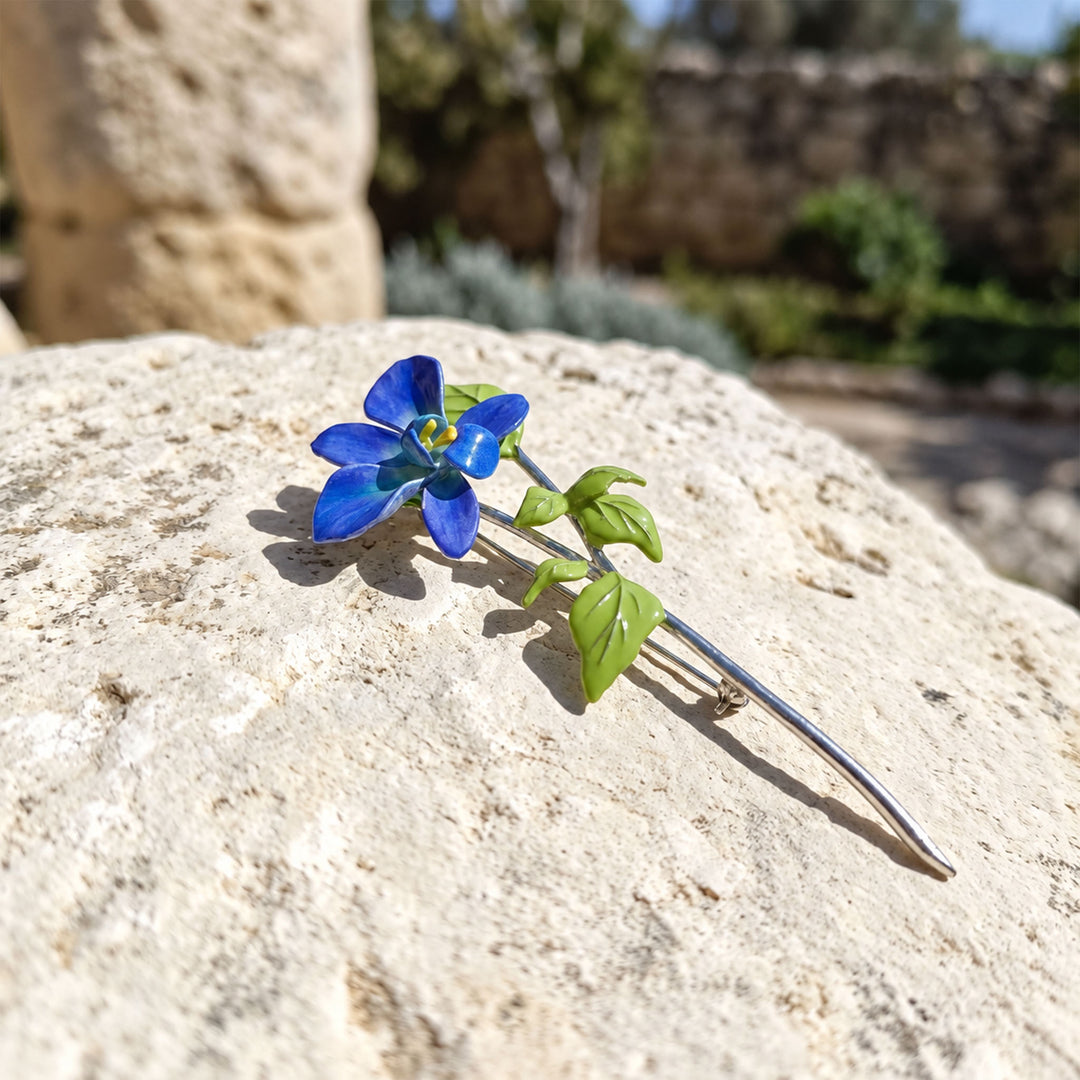 Close-up of a hand-painted blue orchid brooch with green leaves and a long silver-tone stem on textured stone.