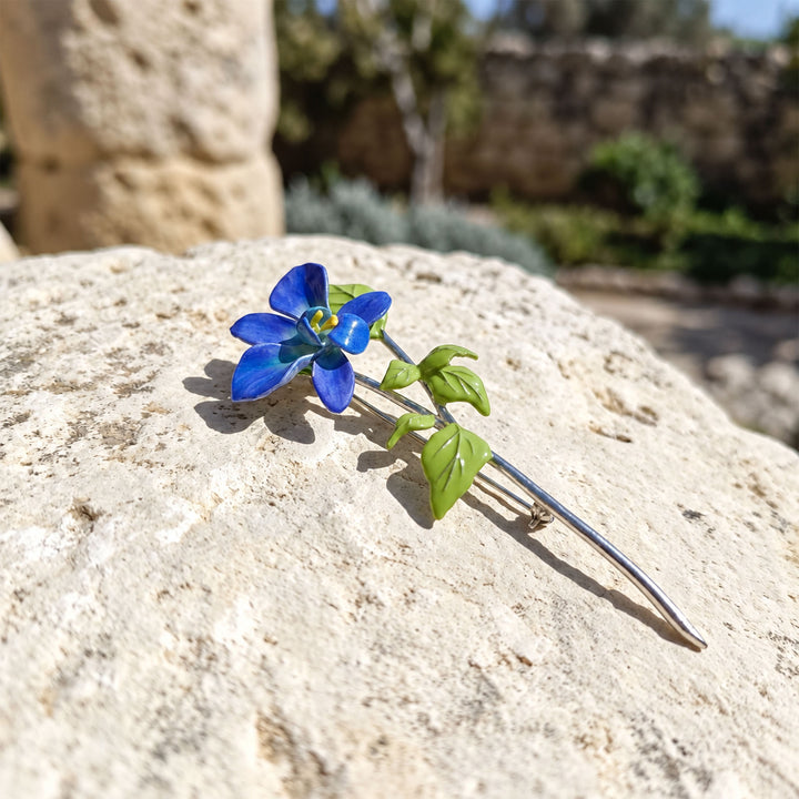 Close-up of a hand-painted blue orchid brooch with green leaves and a long silver-tone stem on textured stone.