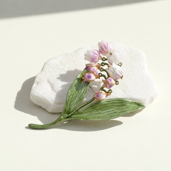 Handcrafted lilac Lily of the Valley floral brooch with white bell-shaped blooms and green leaves on a cream background.