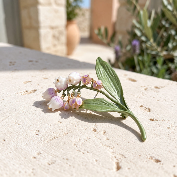 Lilac Lily of the Valley brooch with mother-of-pearl floral details and sculptural green leaves on sunlit pale stone.