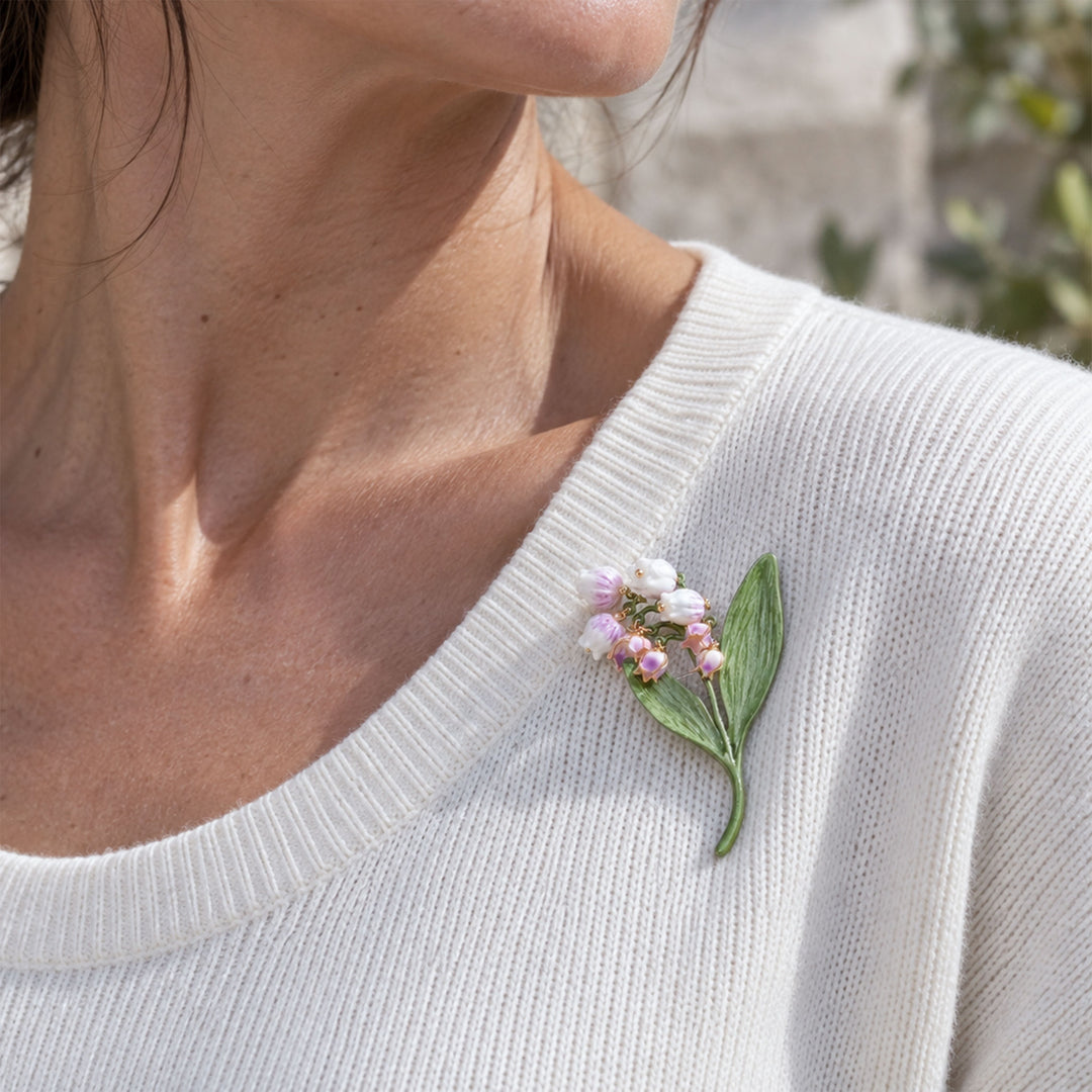 A close-up of a woman wearing a delicate lilac Lily of the Valley brooch with hand-painted flowers and green leaves on the chest of an off-white ribbed knit sweater.