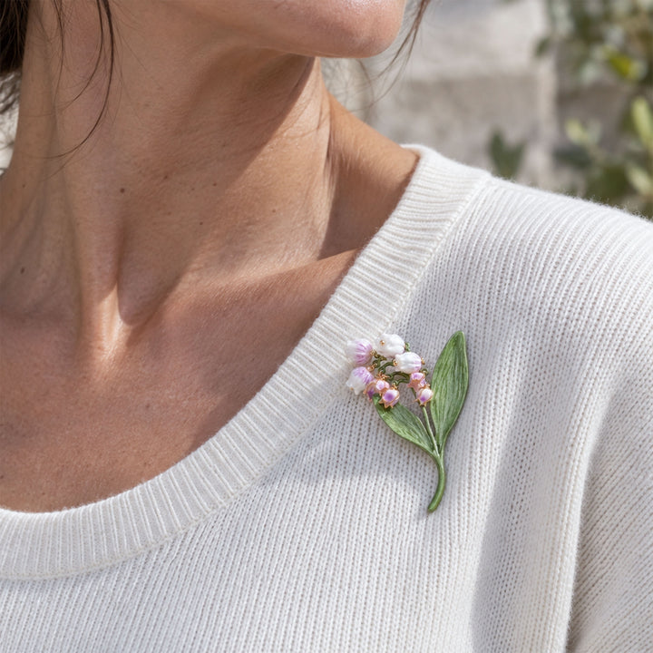A close-up of a woman wearing a delicate lilac Lily of the Valley brooch with hand-painted flowers and green leaves on the chest of an off-white ribbed knit sweater.