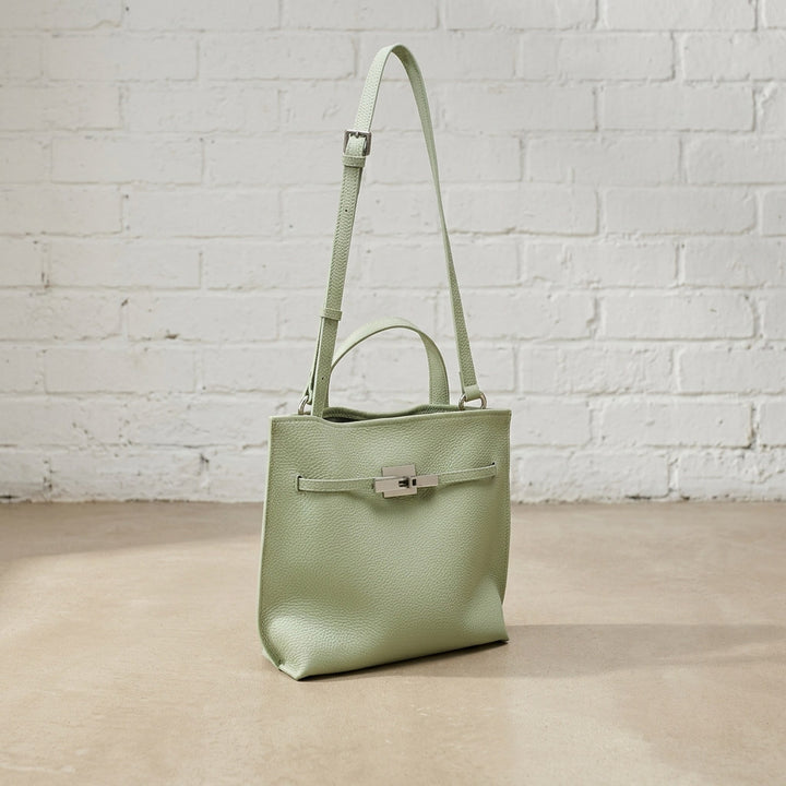 Sage Green Soft Grain Mini Shoulder Bag with pebbled leather texture, top handle, and adjustable shoulder strap against a white brick wall.