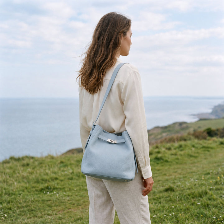Model wearing the Powder Blue Belted Bucket Bag crossbody with a cream outfit against a coastal landscape.