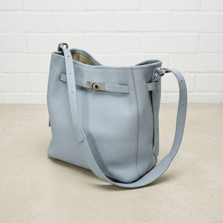 Powder Blue Belted Bucket Bag with textured leather, silver-tone front hardware, and adjustable strap against a white brick wall.