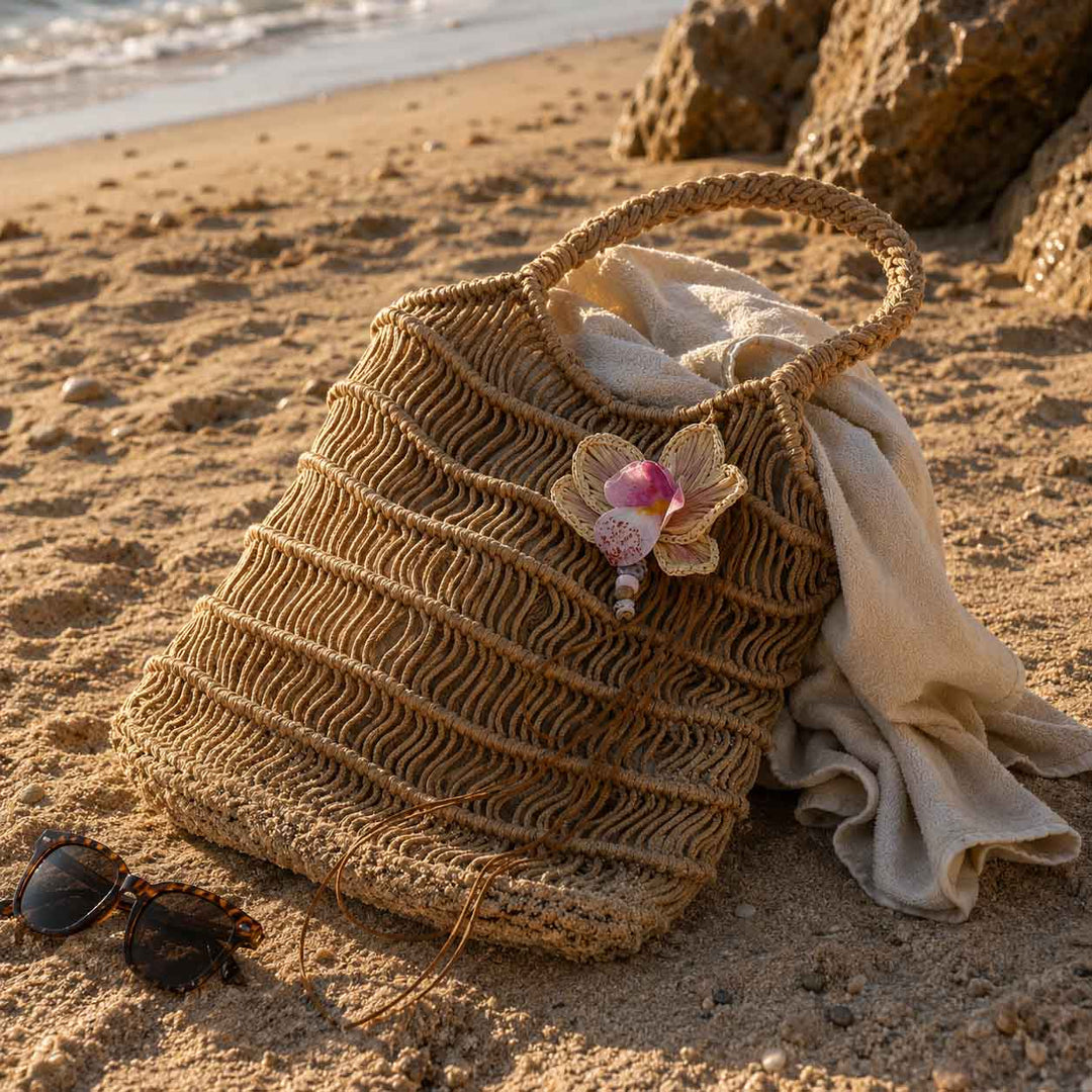 A woven straw beach tote bag sits on a sandy beach next to sunglasses and a cream towel, with rocks and the ocean in the background.