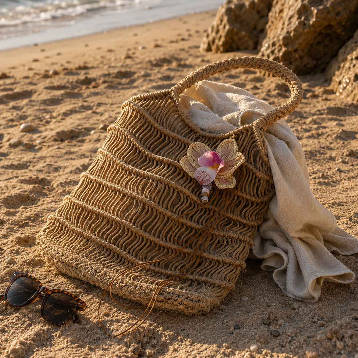 A woven straw beach tote bag sits on a sandy beach next to sunglasses and a cream towel, with rocks and the ocean in the background.