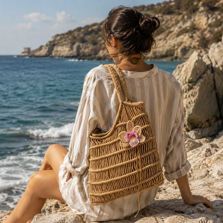 A woman sitting on a rocky beach, viewed from behind, wearing a cream-colored striped shirt and carrying a woven straw tote bag with a pink raffia orchid accent. The background features the blue sea and rocky coastline.