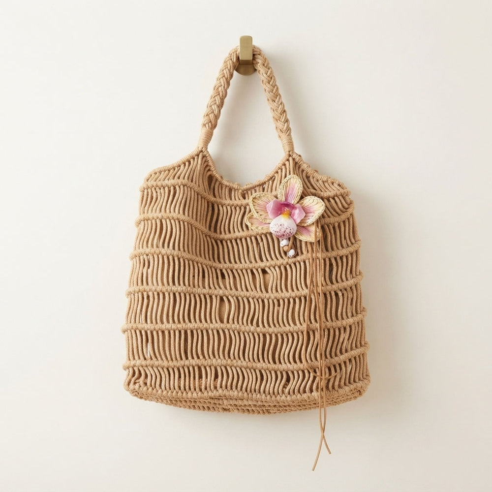 A natural beige woven rope tote bag with braided shoulder straps, decorated with a pink and white orchid flower accent on the front, hanging against a plain off-white wall.