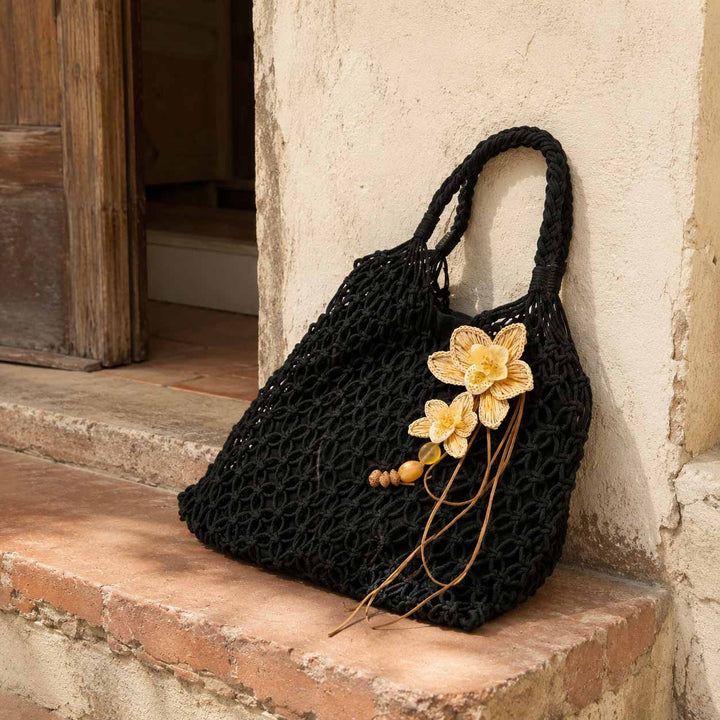 A black crocheted shoulder bag with two top handles, decorated with hand woven raffia orchid and brown wooden beads, resting on a terracotta step against a textured beige wall.