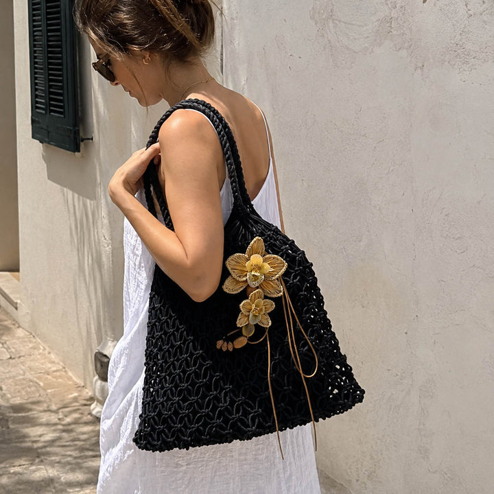 A woman in a white dress carries a large black crocheted shoulder bag with yellow hand woven raffia orchid embellishments, standing against a textured white wall.