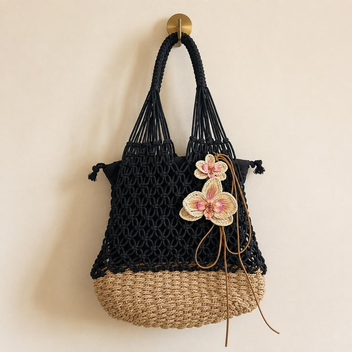A black macrame tote bag with a natural woven straw bottom, decorated with hand weaved raffia orchid flowers, hanging from a gold hook on a beige wall.