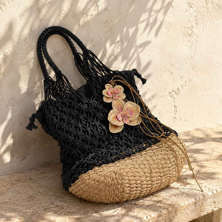 A black crocheted tote bag with a natural beige woven base, decorated with hand weaved raffia orchid resting on a stone ledge against a textured stucco wall.