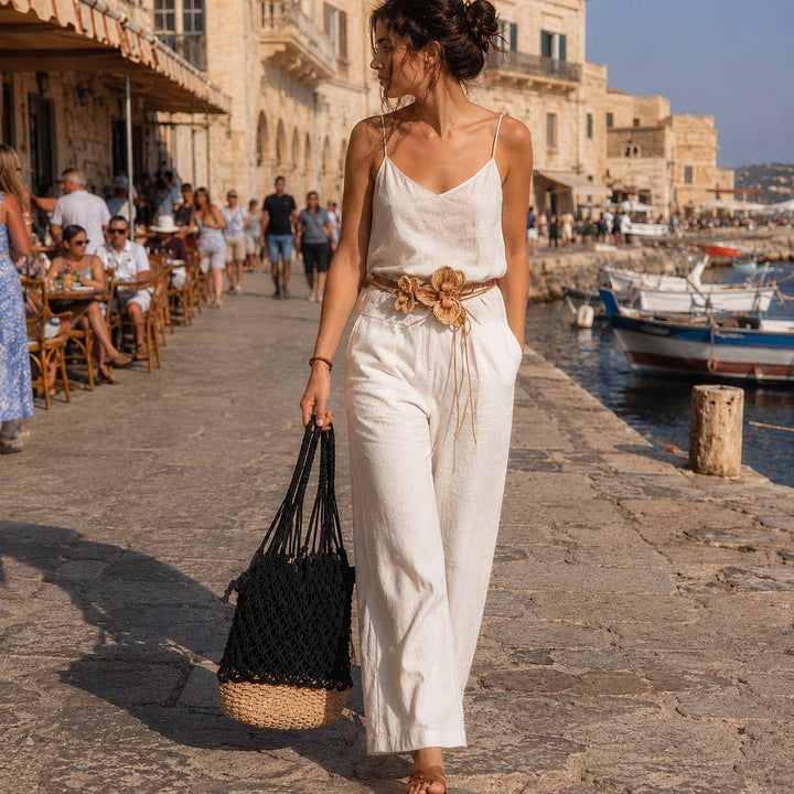 A woman walks along a sunny stone waterfront promenade wearing a white sleeveless jumpsuit with thin straps, a tan woven belt with orchid detail, and carrying a black woven tote bag. Stone buildings, seated people, and boats are visible in the background.
