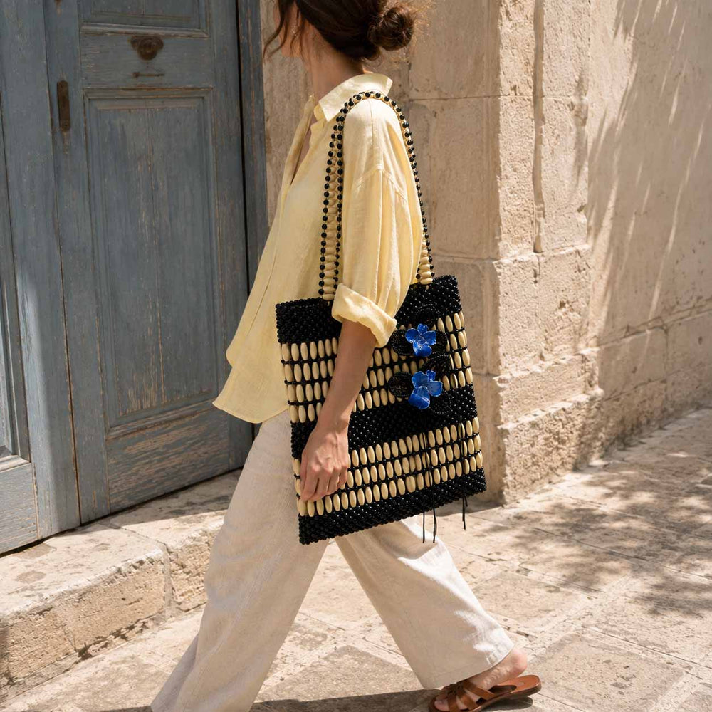A woman walking outdoors beside a stone wall and blue door, carrying a large woven tote bag. The bag is black and cream wooden beads with a repeating geometric pattern and two blue orchid embellishments on the side.