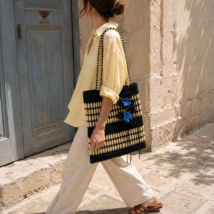 A woman walking outdoors beside a stone wall and blue door, carrying a large woven tote bag. The bag is black and cream wooden beads with a repeating geometric pattern and two blue orchid embellishments on the side.