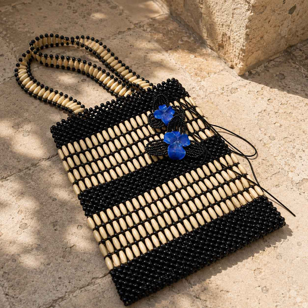 A woven handbag with black and beige wooden beads, featuring t removable blue orchid floral details on the front, resting on a stone surface.