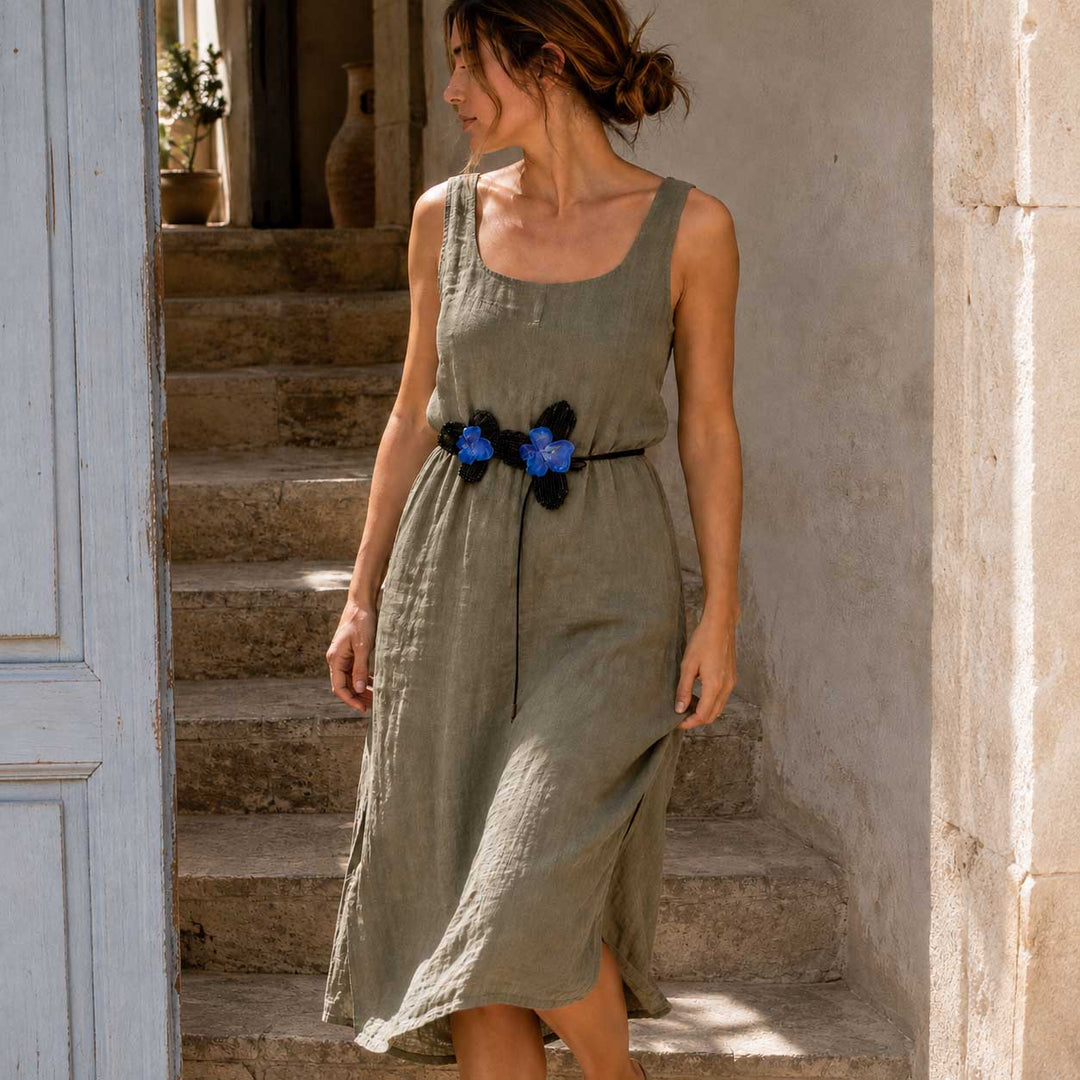 A woman wearing a sleeveless olive green linen midi dress with a black and blue crocheted orchid flower belt, standing on stone steps in a rustic doorway.