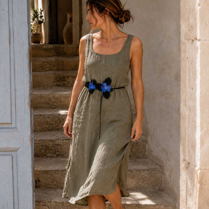 A woman wearing a sleeveless olive green linen midi dress with a black and blue crocheted orchid flower belt, standing on stone steps in a rustic doorway.
