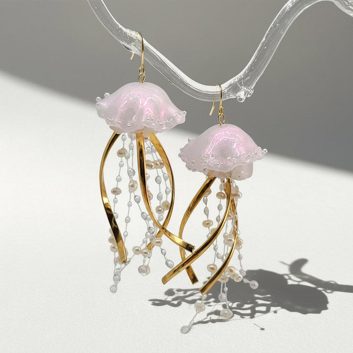 Jellyfish-shaped earrings with pink tops and gold and pearl details on a clear stand.