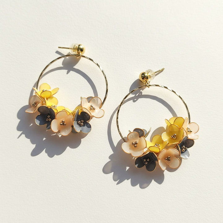 Gold hoop earrings with yellow, cream, and gray enamel flowers, crafted floral design on light background.