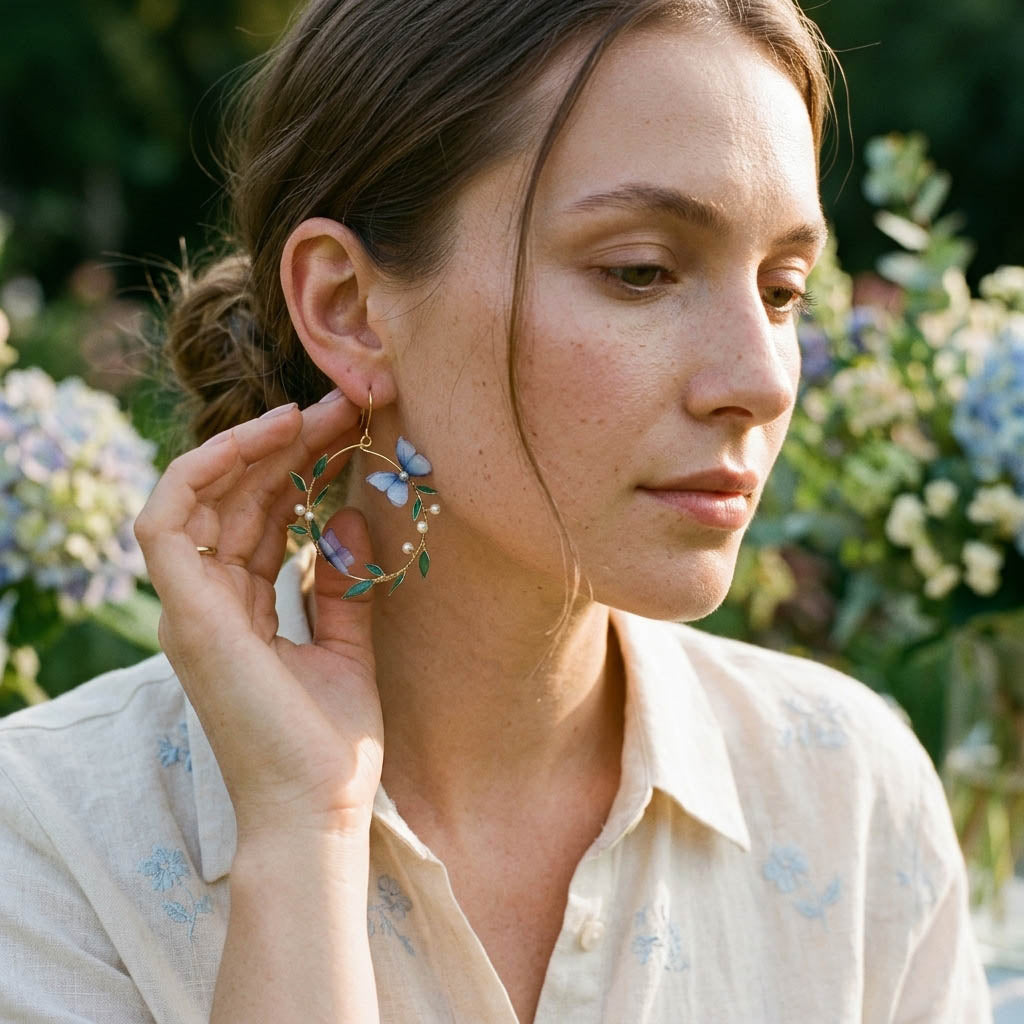 Hand-painted blue floral hoop earrings with green leaf details in gold, worn by model in soft natural garden light
