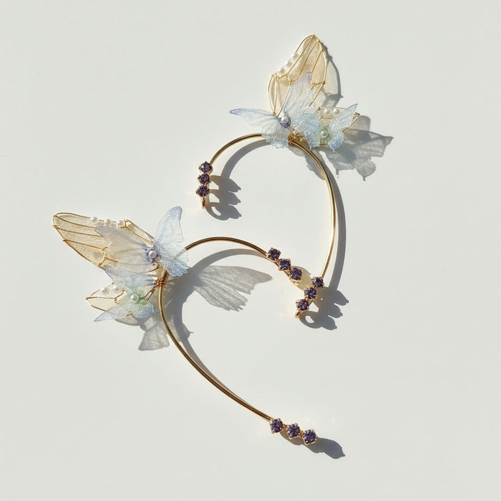 Gold butterfly ear cuffs with translucent wings, pearl accents, and purple crystals on light background.