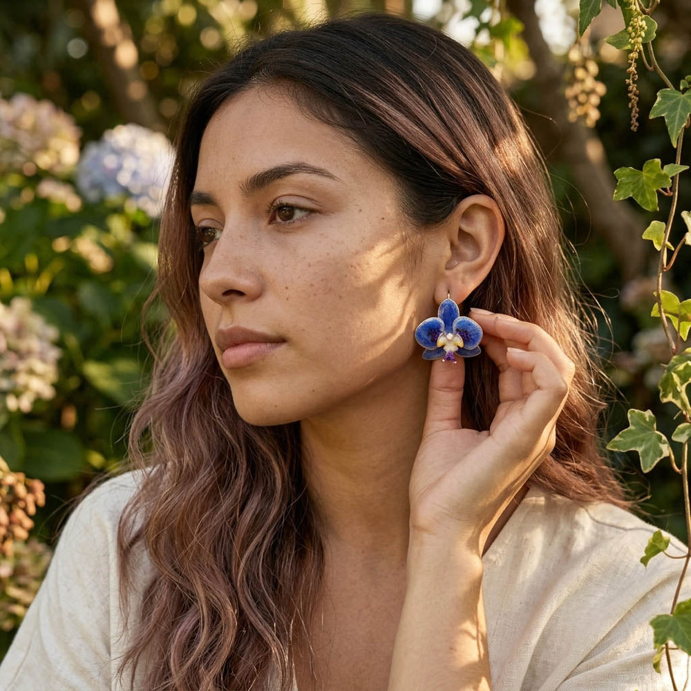 Woman wearing blue orchid earrings outdoors with greenery in the background