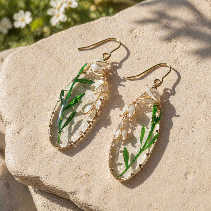 Pair of earrings with green leaves and clear stones on a stone surface with flowers in the background
