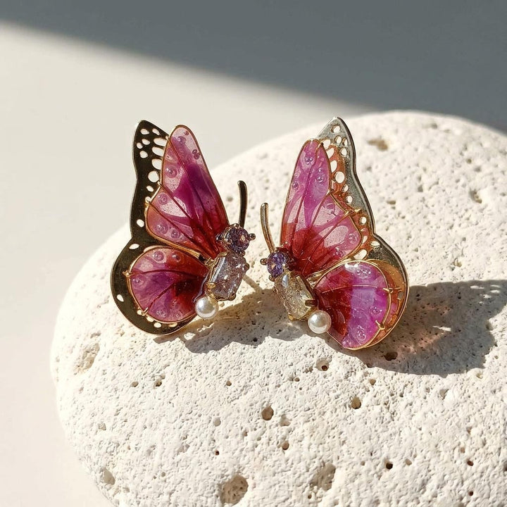 February birthstone butterfly-shaped earrings with pink wings and gold accents on a textured stone surface.