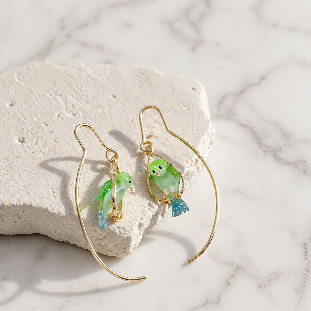 Gold earrings with green and blue bird charms on a marble surface