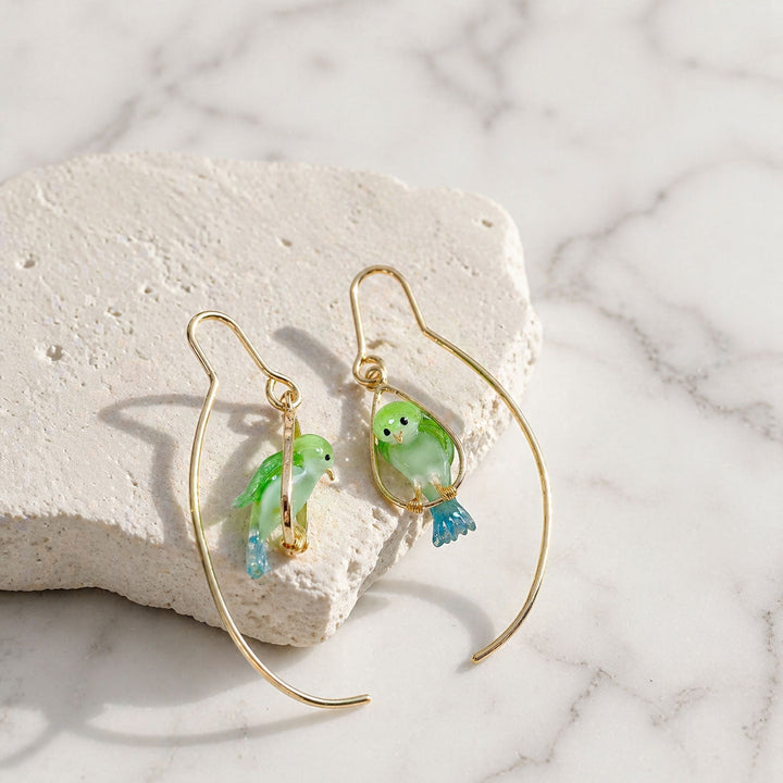 Gold earrings with green and blue bird charms on a marble surface