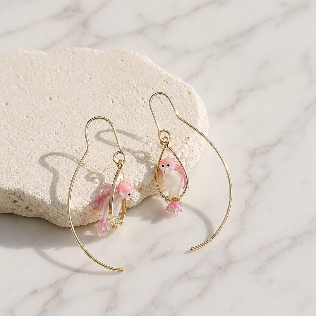 Gold hoop earrings with pink bird charms on a marble surface