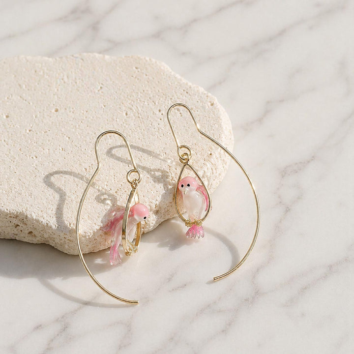 Gold hoop earrings with pink bird charms on a marble surface