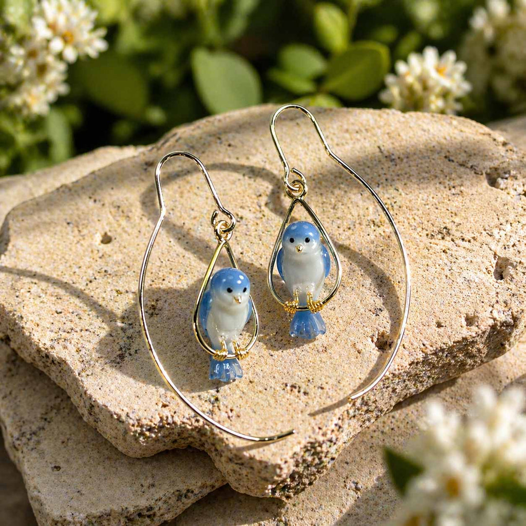 Pair of blue bird-shaped earrings on a stone surface with flowers in the background