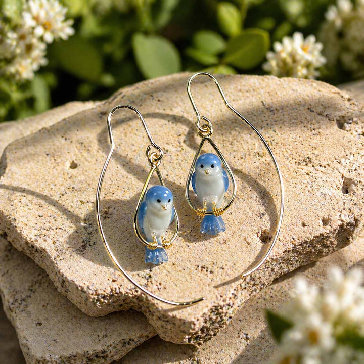 Pair of blue bird-shaped earrings on a stone surface with flowers in the background