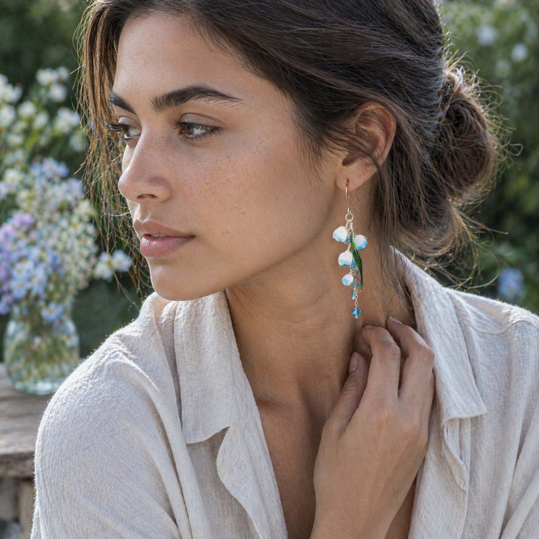 Blue and green lily-of-the-valley drop earrings worn by a woman in a soft outdoor garden portrait, styled with a light ivory linen shirt, natural skin texture, and neutral sunlight, with blurred greenery and pale flowers in the background.