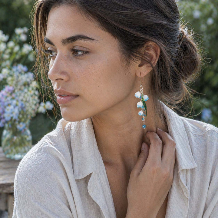 Blue and green lily-of-the-valley drop earrings worn by a woman in a soft outdoor garden portrait, styled with a light ivory linen shirt, natural skin texture, and neutral sunlight, with blurred greenery and pale flowers in the background.