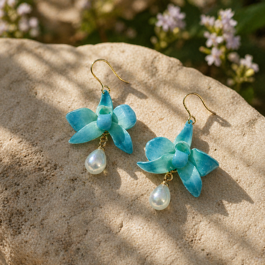 Blue orchid flower-shaped earrings with pearl drops on a stone surface with blurred flowers in the background