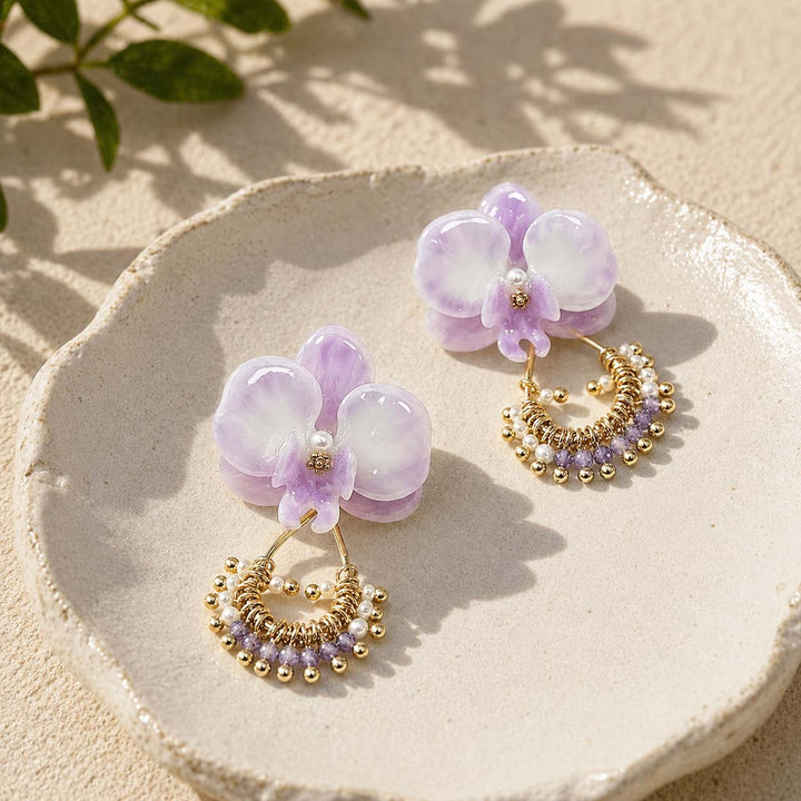 Pair of purple orchid earrings with gold and pearl details on a beige plate.
