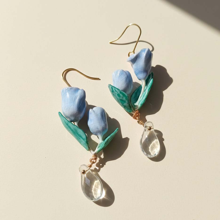 Blue tulip flower drop earrings with green leaves and teardrop crystals, handcrafted resin floral jewelry in sunlight.
